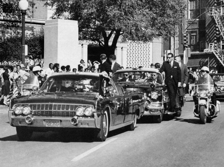 In this Friday, Nov. 22, 1963 file photo, seen through the foreground convertible's windshield, U.S. President John F. Kennedy's hand reaches toward his head within seconds of being fatally shot as first lady Jacqueline Kennedy holds his forearm while the motorcade proceeds along Elm Street past the Texas School Book Depository in Dallas.  